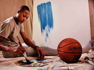 A person is seated on a canvas-covered floor, holding a paintbrush and mixing blue paint on a palette. In front of them is a canvas with a large blue brushstroke. A basketball is placed nearby, and several tubes of paint are scattered on the floor. The setting suggests a blend of art and sports.