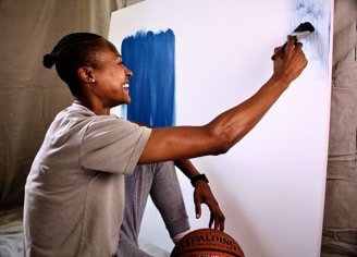 A person with braided hair is sitting on the floor in front of a blank canvas, smiling as they apply blue paint with a brush. They are wearing a light-colored t-shirt and holding a basketball. The background is a neutral fabric drape.
