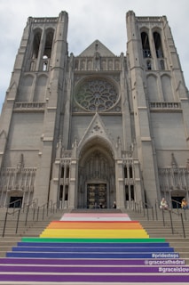 A large, ornate cathedral with a prominent Gothic architectural style, featuring a grand rose window above the entrance. The stairs leading up to the entrance are painted in a rainbow design, symbolizing inclusivity and diversity. People are seen sitting on or near the steps.