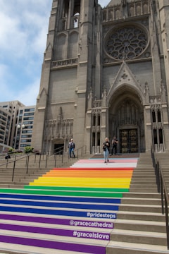 A large Gothic-style cathedral with detailed stone architecture, including a large circular stained glass window above the entrance. The steps leading up to the entrance are painted in vibrant rainbow colors, representing the LGBTQ+ pride flag. There are a few people walking on the steps and at the entrance of the cathedral. Buildings and trees are visible in the background.