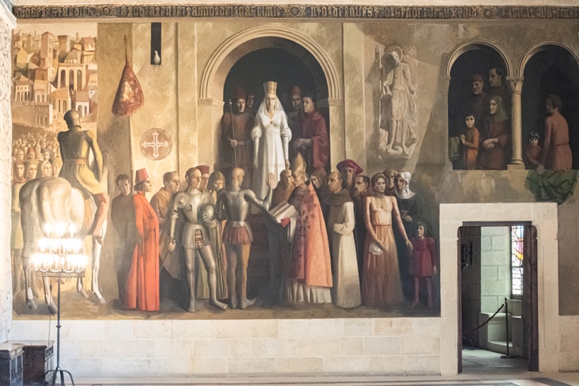 A historical mural featuring various figures dressed in medieval attire, including armored knights, clergymen in religious garments, and a central figure in white robes with a crown suggesting high status. The setting appears to be a grand hall or chamber with intricate architectural details, including arches and sculptures. The scene captures a moment of gathering or ceremony, with a diverse group of people present, possibly indicating an important event or ritual.