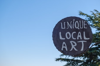 A circular sign displays the words 'Unique Local Art' in white letters against a dark background. The sign is mounted on a metal frame with a clear blue sky in the background and branches of a tree visible to the right.