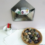 A metal envelope-shaped wall organizer holds several colorful cartoon-themed cards. Below it, a small white container holds red and brown markers next to a few sheets of white paper. A wicker basket contains painted stones with letters on them, arranged neatly.