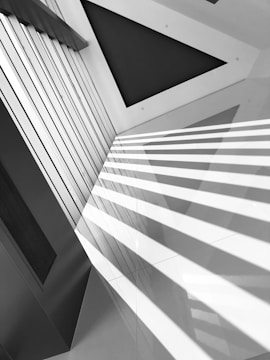 A modern architectural interior featuring clean lines and geometric shapes, with strong diagonal shadows cast across a sleek, monochromatic surface. The composition emphasizes symmetry and contrast within a structured space.