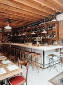A cozy bar setting with a long counter and high stools arranged along it. The bar has a brick wall background adorned with numerous bottles and glassware on shelves. Several pendant lights hang from the wooden ceiling. In the foreground, there are wooden tables and chairs set with menus and glasses.