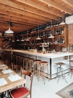 A cozy bar setting with a long counter and high stools arranged along it. The bar has a brick wall background adorned with numerous bottles and glassware on shelves. Several pendant lights hang from the wooden ceiling. In the foreground, there are wooden tables and chairs set with menus and glasses.