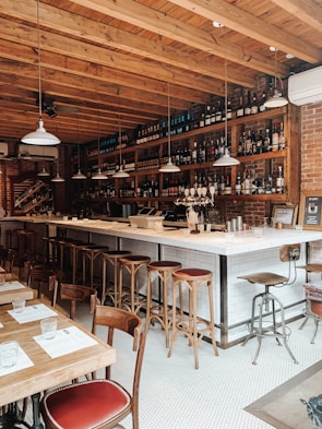 A cozy bar setting with a long counter and high stools arranged along it. The bar has a brick wall background adorned with numerous bottles and glassware on shelves. Several pendant lights hang from the wooden ceiling. In the foreground, there are wooden tables and chairs set with menus and glasses.