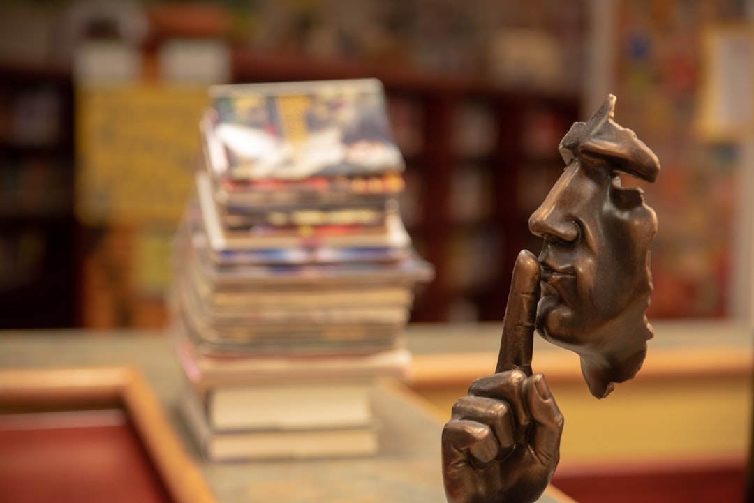 stack of stack of books with sculpture in foreground with a face and finger over the mouth