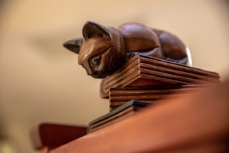 A wooden sculpture of a cat is positioned on top of a stack of books. The craftsmanship highlights the smooth, polished surface and detailed features of the cat, with its ears perked up and eyes focused. The stack of books has visible layers, showcasing the texture of the wood. The background is softly blurred, drawing attention to the intricately carved figure.
