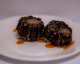 Two dark, chocolatey desserts with a gooey caramel topping are placed on a white plate. Caramel sauce is drizzled over the plate, adding a rich, indulgent touch to the presentation.