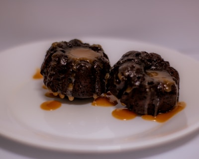 Two dark, chocolatey desserts with a gooey caramel topping are placed on a white plate. Caramel sauce is drizzled over the plate, adding a rich, indulgent touch to the presentation.