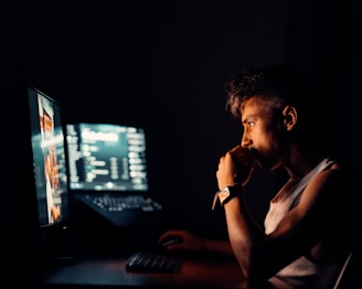 A person is sitting in front of two computer monitors in a dark room, intently looking at the screen with their hand on their chin. They are wearing a sleeveless shirt and a watch.