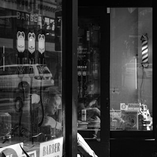 A black and white image of a barber shop window featuring various stickers, including a Bitcoin symbol and notices about WiFi availability. A classic barber pole is mounted near the entrance. The reflections on the glass show street activity and the interior reveals a man sitting inside, looking down at his phone.