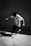 A person is captured mid-motion, seemingly jumping or performing a trick with a skateboard nearby. The setting appears to be urban, with a concrete wall and floor. The lighting creates a high-contrast, black-and-white effect that accentuates the movement and shadows.