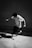 A person is captured mid-motion, seemingly jumping or performing a trick with a skateboard nearby. The setting appears to be urban, with a concrete wall and floor. The lighting creates a high-contrast, black-and-white effect that accentuates the movement and shadows.