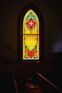 A beautifully colored stained glass window with vibrant patterns featuring red, blue, yellow, and green geometric shapes. The window is set within a darkened room, casting subtle light onto a pew. Below the window, the name 'Rev. A. C. Shriver' is inscribed.