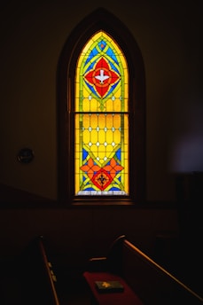 A beautifully colored stained glass window with vibrant patterns featuring red, blue, yellow, and green geometric shapes. The window is set within a darkened room, casting subtle light onto a pew. Below the window, the name 'Rev. A. C. Shriver' is inscribed.