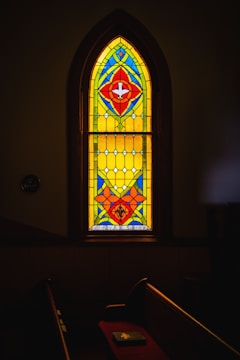 A beautifully colored stained glass window with vibrant patterns featuring red, blue, yellow, and green geometric shapes. The window is set within a darkened room, casting subtle light onto a pew. Below the window, the name 'Rev. A. C. Shriver' is inscribed.