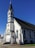 A white, wooden church building with a tall steeple featuring a cross at the top. The structure has dark-colored roofing and several arched windows with stained glass. A ramp and steps lead up to the entrance, which is located at the front of the building.