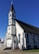 A white, wooden church building with a tall steeple featuring a cross at the top. The structure has dark-colored roofing and several arched windows with stained glass. A ramp and steps lead up to the entrance, which is located at the front of the building.