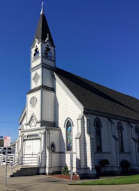 A white, wooden church building with a tall steeple featuring a cross at the top. The structure has dark-colored roofing and several arched windows with stained glass. A ramp and steps lead up to the entrance, which is located at the front of the building.