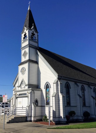 A white, wooden church building with a tall steeple featuring a cross at the top. The structure has dark-colored roofing and several arched windows with stained glass. A ramp and steps lead up to the entrance, which is located at the front of the building.