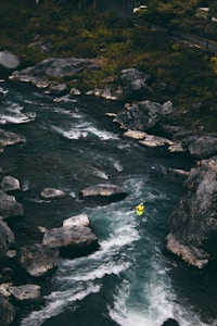 A person in a bright yellow kayak navigates through a flowing river with rapids. The water is flanked by large rocks and surrounded by dense, green foliage. The scene captures a sense of adventure and nature's rugged beauty.