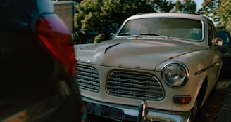 A classic, vintage Volvo car is parked on the street. The front grill and headlights are prominently visible, surrounded by a slightly worn and chipped white exterior. Parked in a line of cars, the environment includes leafy trees and a residential backdrop. The lighting suggests a setting sun or an overcast day.