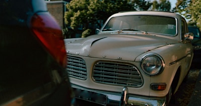 A classic, vintage Volvo car is parked on the street. The front grill and headlights are prominently visible, surrounded by a slightly worn and chipped white exterior. Parked in a line of cars, the environment includes leafy trees and a residential backdrop. The lighting suggests a setting sun or an overcast day.