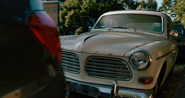A classic, vintage Volvo car is parked on the street. The front grill and headlights are prominently visible, surrounded by a slightly worn and chipped white exterior. Parked in a line of cars, the environment includes leafy trees and a residential backdrop. The lighting suggests a setting sun or an overcast day.