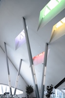 Modern architectural interior featuring a high ceiling with multiple skylights in vibrant colors, including green, yellow, red, and blue. There are several angled steel beams and minimalist decor. Potted plants are positioned near the windows, enhancing the open and airy atmosphere.