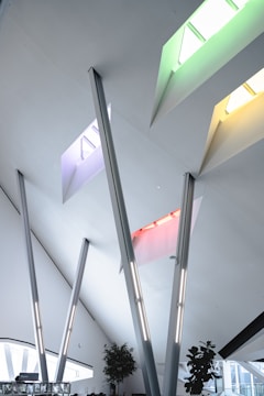 Modern architectural interior featuring a high ceiling with multiple skylights in vibrant colors, including green, yellow, red, and blue. There are several angled steel beams and minimalist decor. Potted plants are positioned near the windows, enhancing the open and airy atmosphere.