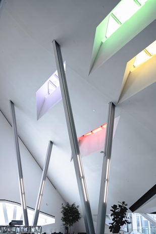 Modern architectural interior featuring a high ceiling with multiple skylights in vibrant colors, including green, yellow, red, and blue. There are several angled steel beams and minimalist decor. Potted plants are positioned near the windows, enhancing the open and airy atmosphere.