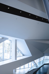 The image depicts a modern architectural interior with sharp geometric lines, large windows allowing ample natural light, and a minimalist aesthetic. The space features white walls, a sleek black beam with inset circle lights, and a railing along a stairway or hallway.