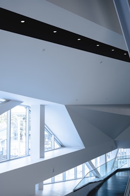 The image depicts a modern architectural interior with sharp geometric lines, large windows allowing ample natural light, and a minimalist aesthetic. The space features white walls, a sleek black beam with inset circle lights, and a railing along a stairway or hallway.