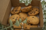 A box containing several chocolate chip cookies, partially surrounded by green leaves. The cookies are golden-brown with visible dark chocolate chips and are placed on a sheet of paper inside the box.