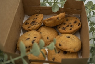 A box containing several chocolate chip cookies, partially surrounded by green leaves. The cookies are golden-brown with visible dark chocolate chips and are placed on a sheet of paper inside the box.