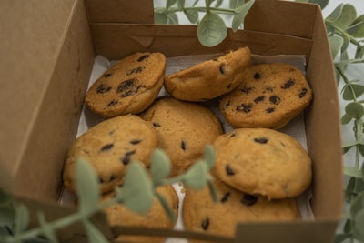 A box containing several chocolate chip cookies, partially surrounded by green leaves. The cookies are golden-brown with visible dark chocolate chips and are placed on a sheet of paper inside the box.