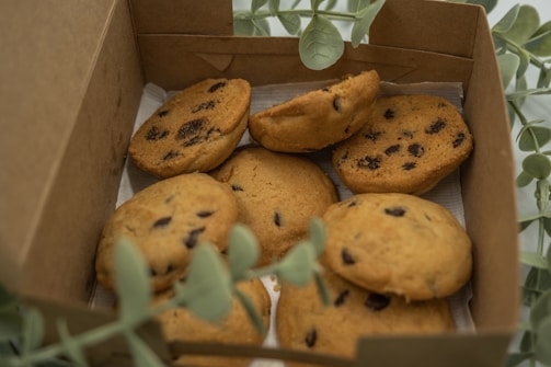 A box containing several chocolate chip cookies, partially surrounded by green leaves. The cookies are golden-brown with visible dark chocolate chips and are placed on a sheet of paper inside the box.