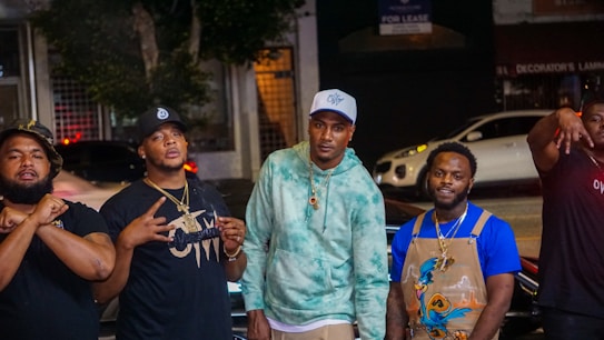 A group of five men stand on a street, posing for a photo. They are in casual clothing, with some wearing hats and jewelry. The background includes parked cars, a 'For Lease' sign, and trees, suggesting an urban environment at night.