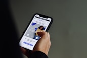 A person is holding a smartphone with both hands, browsing a website. The screen displays a webpage with text in Portuguese and features an image of a smiling person against a wall. The background is a neutral color, focusing attention on the phone and its content.