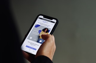 A person is holding a smartphone with both hands, browsing a website. The screen displays a webpage with text in Portuguese and features an image of a smiling person against a wall. The background is a neutral color, focusing attention on the phone and its content.