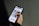 A person is holding a smartphone with both hands, browsing a website. The screen displays a webpage with text in Portuguese and features an image of a smiling person against a wall. The background is a neutral color, focusing attention on the phone and its content.