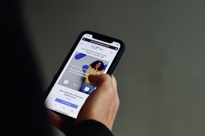 A person is holding a smartphone with both hands, browsing a website. The screen displays a webpage with text in Portuguese and features an image of a smiling person against a wall. The background is a neutral color, focusing attention on the phone and its content.
