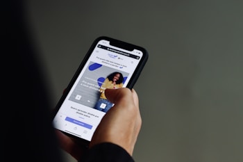 A person is holding a smartphone with both hands, browsing a website. The screen displays a webpage with text in Portuguese and features an image of a smiling person against a wall. The background is a neutral color, focusing attention on the phone and its content.