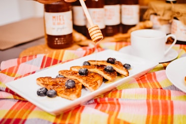 A white rectangular plate is filled with slices of toasted bread topped with fresh blueberries. Honey is being drizzled on the bread from a wooden honey dipper. In the background, jars of honey are visible, along with a white cup near the plate. The setting rests on a colorful, striped cloth, giving the scene a warm and inviting look.