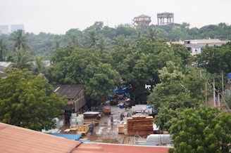 Aerial view of an industrial area surrounded by dense green trees. The scene includes stacked wooden logs, metal sheets, and various materials on the ground. There are several buildings with tin roofs and water towers visible in the background. Utility poles and wires run along the area, suggesting an industrial or commercial setting.