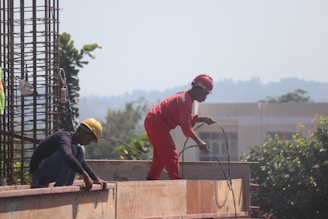 Two construction workers are engaged in building work. One worker in red overalls and a red helmet is standing, holding wires, while the other, wearing a yellow helmet, is crouched down, working on a wooden structure. Rebar and trees can be seen in the background.