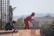 Two construction workers are engaged in building work. One worker in red overalls and a red helmet is standing, holding wires, while the other, wearing a yellow helmet, is crouched down, working on a wooden structure. Rebar and trees can be seen in the background.
