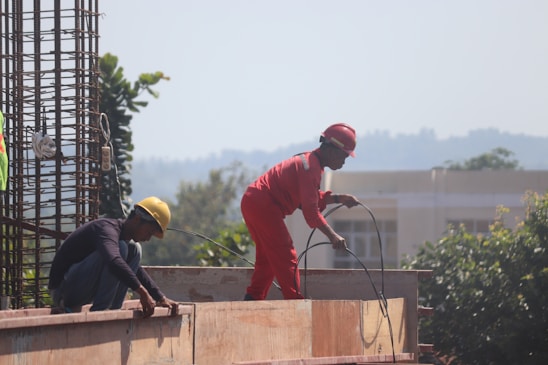 Two construction workers are engaged in building work. One worker in red overalls and a red helmet is standing, holding wires, while the other, wearing a yellow helmet, is crouched down, working on a wooden structure. Rebar and trees can be seen in the background.